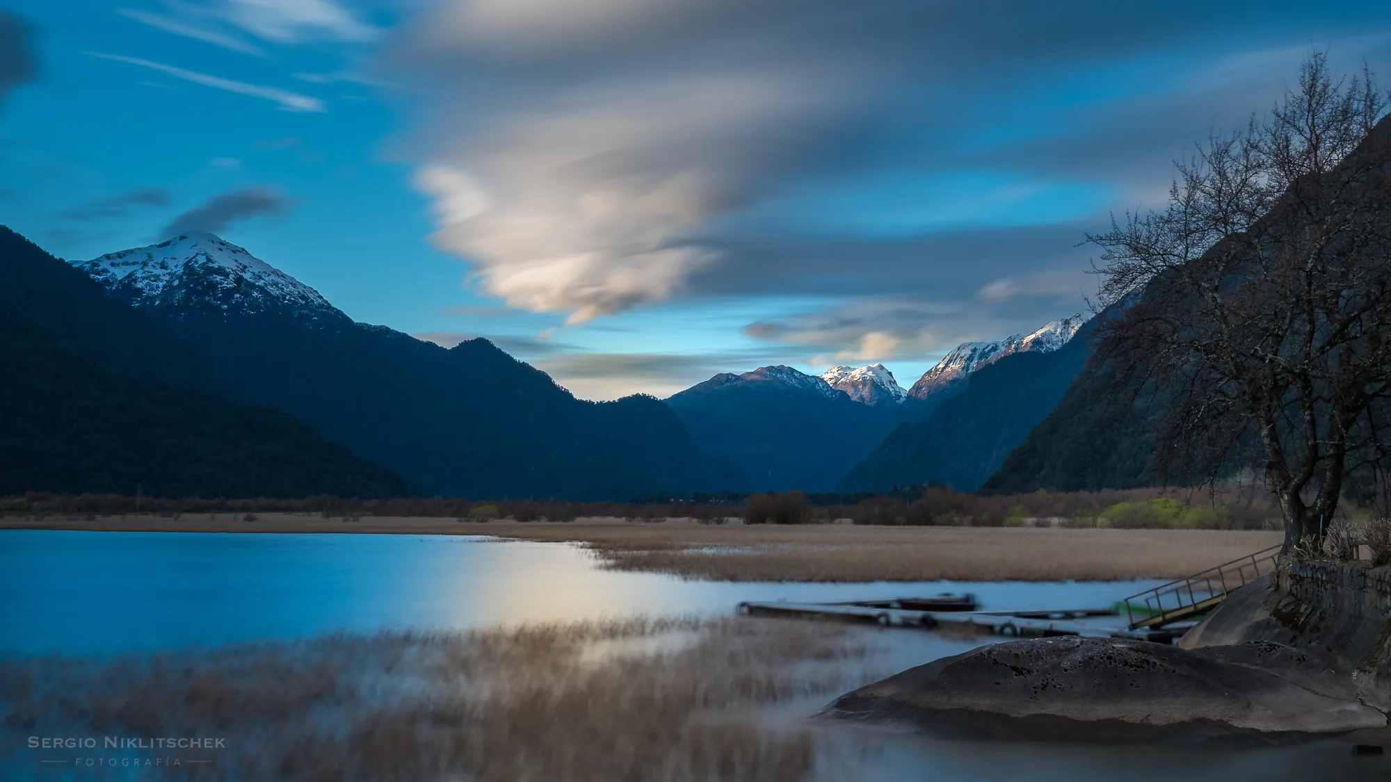 Atardecer de larga exposición sobre el agua, los juncos y las montañas nevadas de Peulla