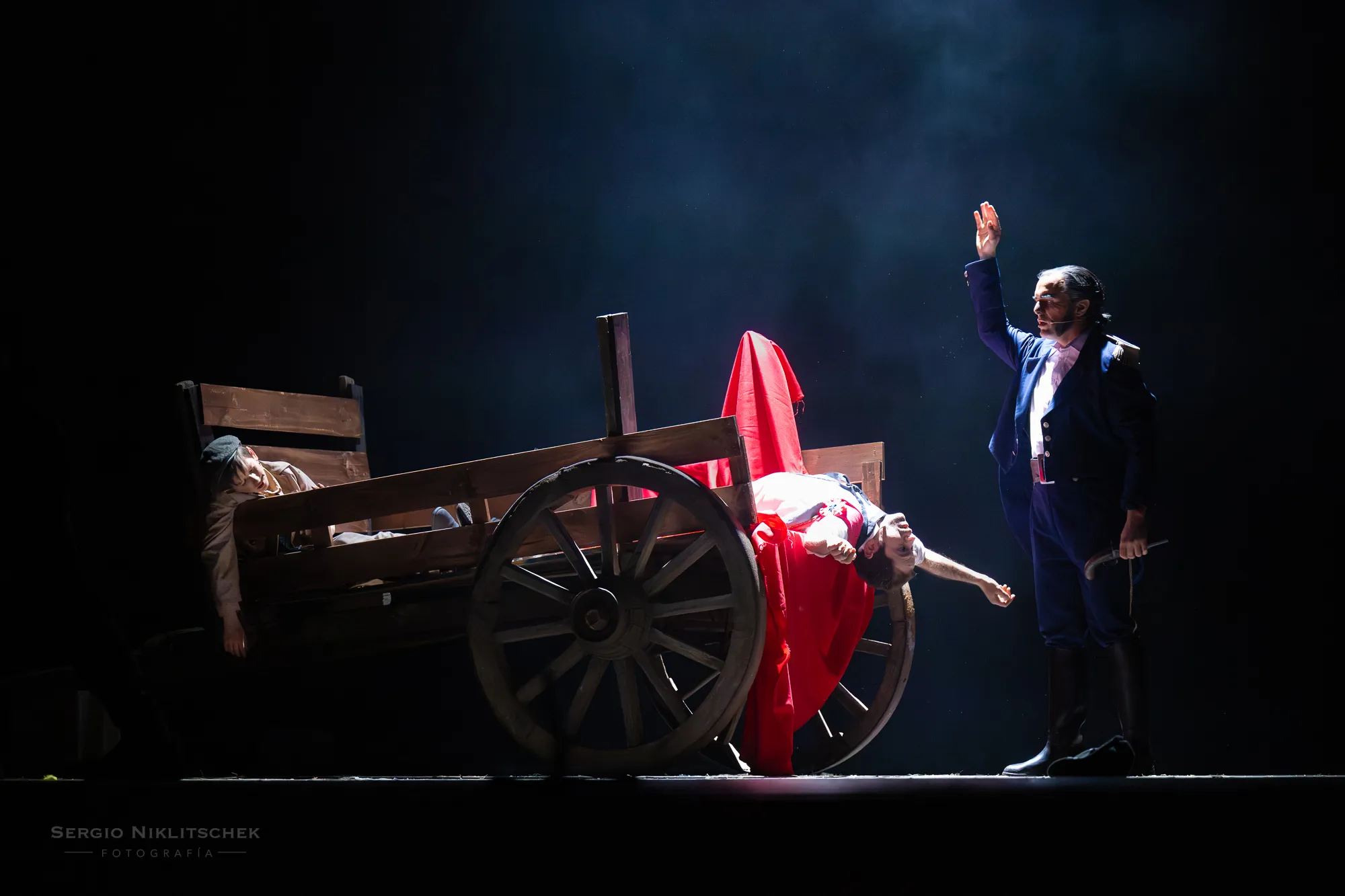 Escena teatral de Los miserables: un actor de uniforme alza la mano junto a una carreta en penumbra