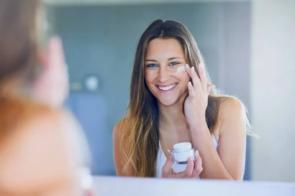 Mujer sonriente aplicándose una crema en la mejilla frente al espejo del baño