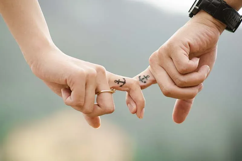 Dos manos entrelazadas por los dedos índice, cada una con un pequeño tatuaje de ancla